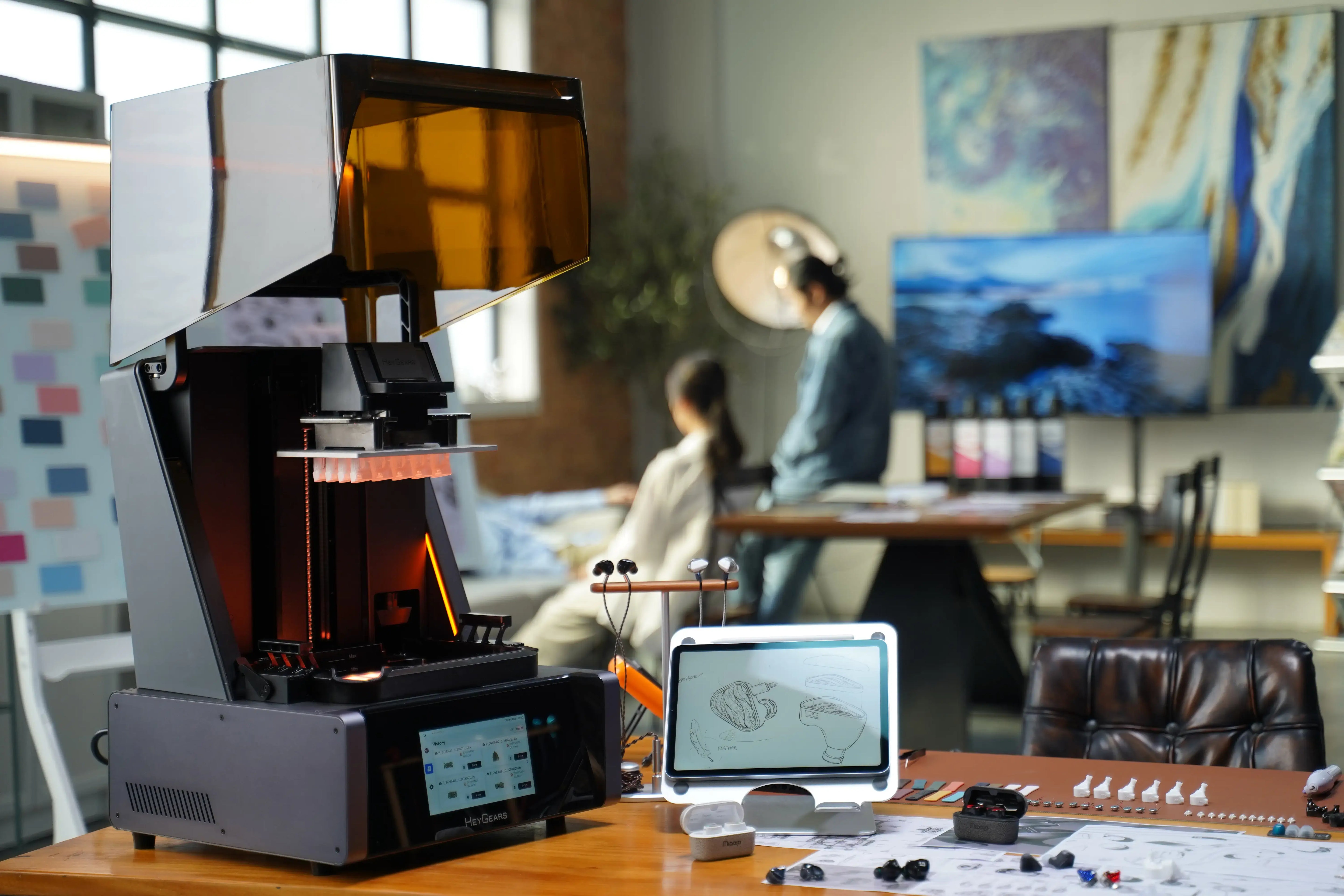 Desktop resin 3D printer with amber cover beside design sketches and printed prototypes in a studio workspace.