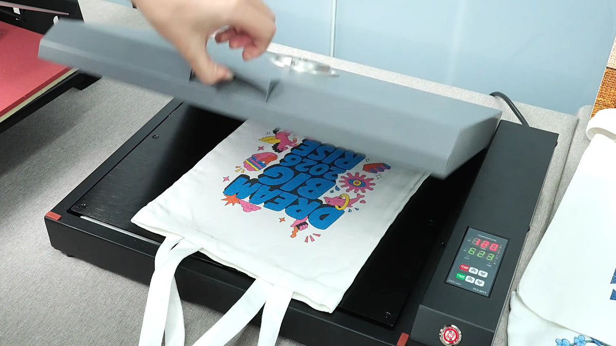 User lifting a heat press above a white tote bag with a colorful DTG printed design during the curing process.